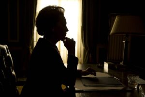 Silhouetted woman at a desk, chin resting on hand, pondering in a dimly lit room with a bright window behind her.