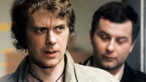 Two men in a tense moment: the foreground man looks worried or angry, while the other stands behind him with eyes closed or looking down.