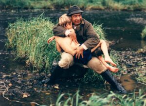 Man wearing a dark hat hugs a shirtless child by a shallow, rocky riverbank with tall grass nearby.
