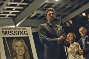 Man in a blazer speaks into a microphone at a missing-person press event beside a large 'MISSING' poster of a smiling woman with the date Thursday July 05, 2012.