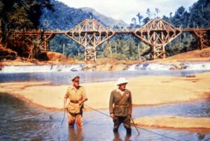 Two soldiers wading in a shallow river with a large wooden truss bridge under construction behind them in a forested, hilly area.