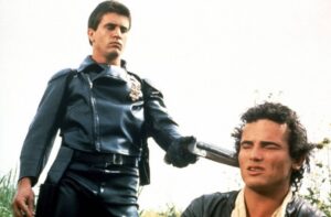 A man in a dark leather police uniform stands over a kneeling man, gripping a baton against his shoulder or neck area, in an outdoor setting.