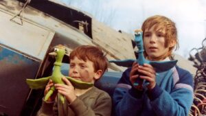 Two children hold colorful dragon hand puppets in front of a damaged vehicle, a green dragon and a blue winged dragon.