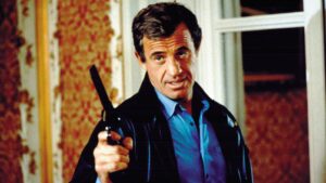 Man wearing a blue shirt and black jacket points a handgun at the camera from indoors, appearing to threaten the viewer.