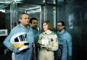 Group of astronauts in blue suits with a woman in a beige suit standing near a glass door, helmet in hand outer edge left.