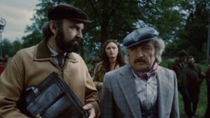 Two men in vintage outfits stand outdoors; the bearded man carries a large leather briefcase, while the other wears a gray cap.