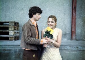 A man and a woman stand together outdoors; she holds a bouquet of yellow flowers and smiles at him.