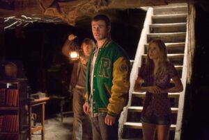 Three people stand in a dim, attic-like room; a man in a green and yellow varsity jacket is foreground, woman by the stairs to the right, and another man holds a lantern in the background.