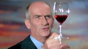Older man in a suit raising a glass of red wine in a toast, looking at the camera.