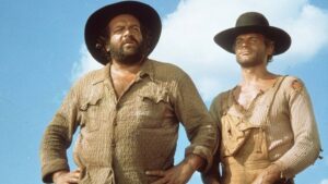Two rugged men in wide-brim hats stand side by side against a blue sky, wearing worn, patched shirts in a desert-like setting.