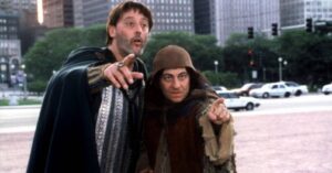 Two men in medieval-style costumes point at something off-camera in a city square.