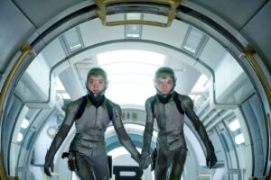Two astronauts in gray suits walk through a futuristic spaceship corridor hand in hand, wearing helmets and green visors.