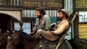Two cowboys ride horses in a dusty Western town, with a weathered storefront sign reading 'BOOTS SHOES' in the background.