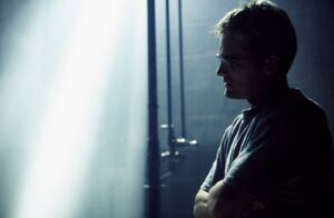 Man in profile with glasses, standing with arms crossed beside a cool blue-lit wall.