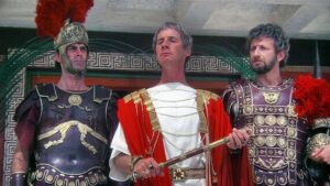 Three men in Roman-inspired armor stand together, the center figure in a white tunic with a red cloak and ceremonial staff flanked by ornate bronze-armored companions.