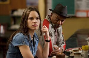 Two people sit at a diner table; a serious-looking woman in a denim shirt holds a napkin, while an older man in a hat sits beside her and looks at her thoughtfully, with a cupcake and coffee cups on the table in front of them.