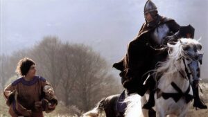 Knight in armor on a white horse rides toward a man in a brown medieval tunic in an open field.