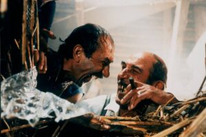 Two men amid debris share a laugh, one holding a cigarette while they cling to rubble and a torn netting in a rescue scene.