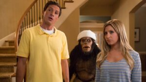 Man in a yellow polo and a woman in a blue striped shirt stand in a home's foyer, a bandaged gorilla behind them looking surprised.
