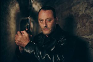 Man in a black leather jacket grips a pistol in a dim tunnel, staring intently at the camera.