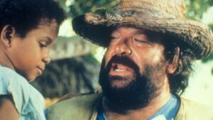 Bearded man in a straw hat speaks softly to a young child outdoors in sunlight.