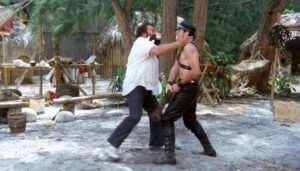 Two men fighting in a rustic outdoor camp, one grabbing the other by the collar as they clash.
