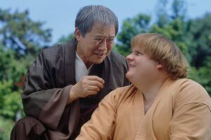 Two martial artists converse outdoors, the older man leaning in with a serious expression toward the younger man in a tan gi.