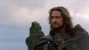 Rugged medieval warrior with long hair and beard in leather armor and cloak, standing by a rocky shore.