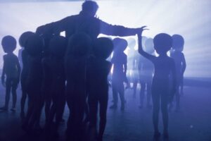 Silhouetted crowd facing a speaker on a raised platform in a hazy blue-lit room.