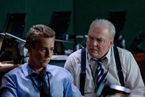 Two men in a dim control room or office, one in a blue shirt and tie looks down, while the older man in a white shirt with suspenders speaks beside him.