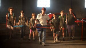 Youth street hockey coach leads a group of kids indoors, each holding hockey sticks, in a dim gym environment.