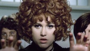 Close-up of a woman with red, curly hair looking at the camera, hands reaching toward the viewer in the foreground.
