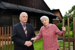 Two elderly people stand outside a dark wooden cottage, a man in a suit and tie beside a woman in a pink knitted sweater near a wooden fence.