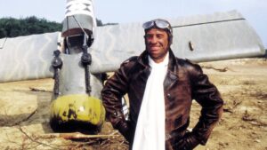 Man in a brown leather jacket and goggles stands beside a crashed helicopter in a dusty, open field.