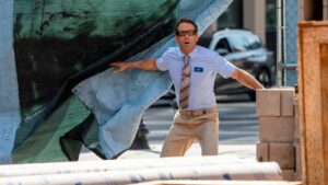 Man in a light blue shirt and striped tie pulls back a large tarp at a construction site, with a surprised expression and a name tag reading 'GUY' on his chest.