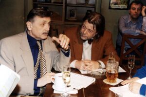 Three men seated at a bar/restaurant table; two in foreground discuss seriously while the third leans back in the background, drinks on the table.