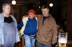 Three middle-aged men stand with arms around each other in a dimly lit pub, a beer mug visible on the table in the foreground.