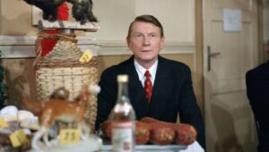 Man in a dark suit and red tie sits at a market stall with sausages and baked goods on the table; a large woven basket and a bottle are visible in the foreground.