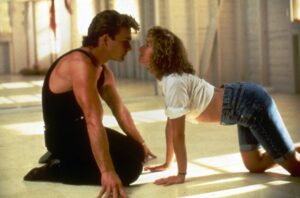 A man in a black sleeveless shirt kneels on the floor facing a woman who is on her hands and knees, their faces inches apart in a kiss-ready pose.