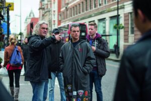 Man with gray hair points as his colleagues listen on a city street; a pink bus is in the background.