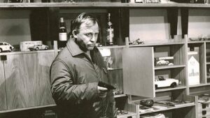 Man in a jacket points a handgun while standing among shelves filled with toy cars and bottles in a shop-like setting, with a serious expression.