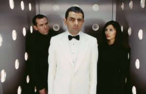 Three people stand in a metallic elevator with circular lights; man in a white tuxedo in the center, flanked by a man and a woman in black behind him.