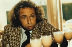 Man with curly hair in a houndstooth blazer toasts with foamy beers toward the camera.