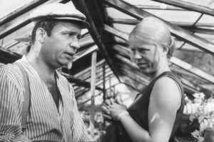 Man in a striped shirt and cap speaks with a blonde woman in a sleeveless dress, their hands clasped as they converse beside a wooden framework.