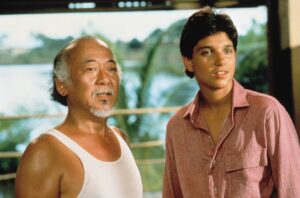 Two men stand side by side outdoors, an older man with a white tank top and gray beard beside a younger man in a pink shirt, tropical greenery in the background.