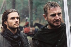 Two men in dark cloaks in a snowy forest, one holding a shiny sword, looking off to the side.