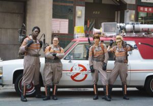 Four Ghostbusters in beige jumpsuits with proton packs pose beside a vintage white and red Ghostbusters car displaying the iconic logo on the door.