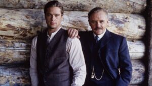 Two men in vintage suits pose in front of a rough log wall, with the older man resting a hand on the younger man's shoulder.