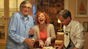 Three friends celebrate around a cake in a cozy dining room; the woman in the center laughs while the men prepare dessert.