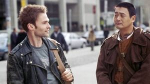Two men walk and chat on a busy city street; the man on the left wears a gray shirt and black leather jacket with a shoulder bag, the man on the right wears a brown coat with a crossbody strap.